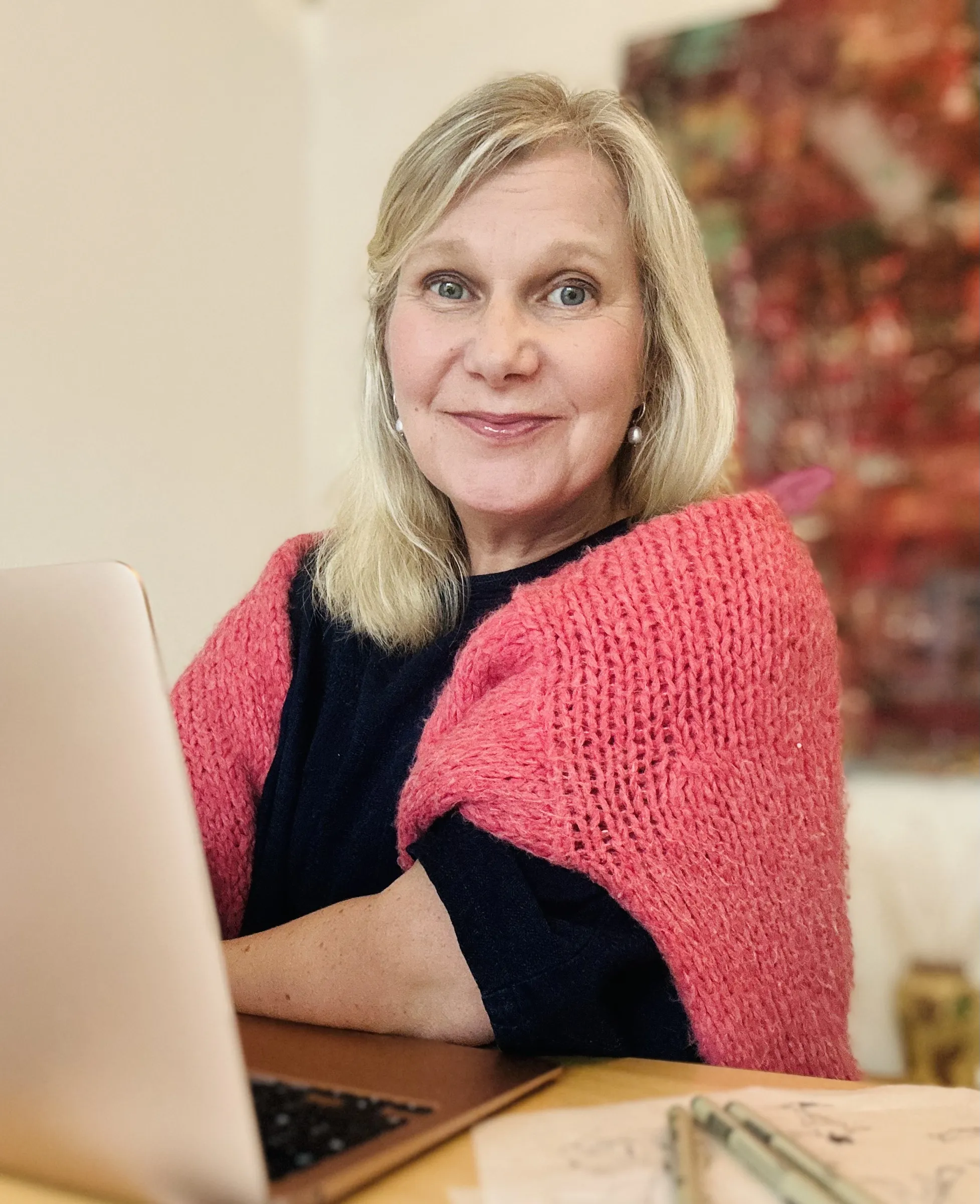 Nainen hymyilee kameralle, hänellä on vaaleat hiukset, vaatteet ovat tummansiniset ja päällä on korallinpunainen neule. Taustalla on abstrakti maalaus ja valo on pehmeää.