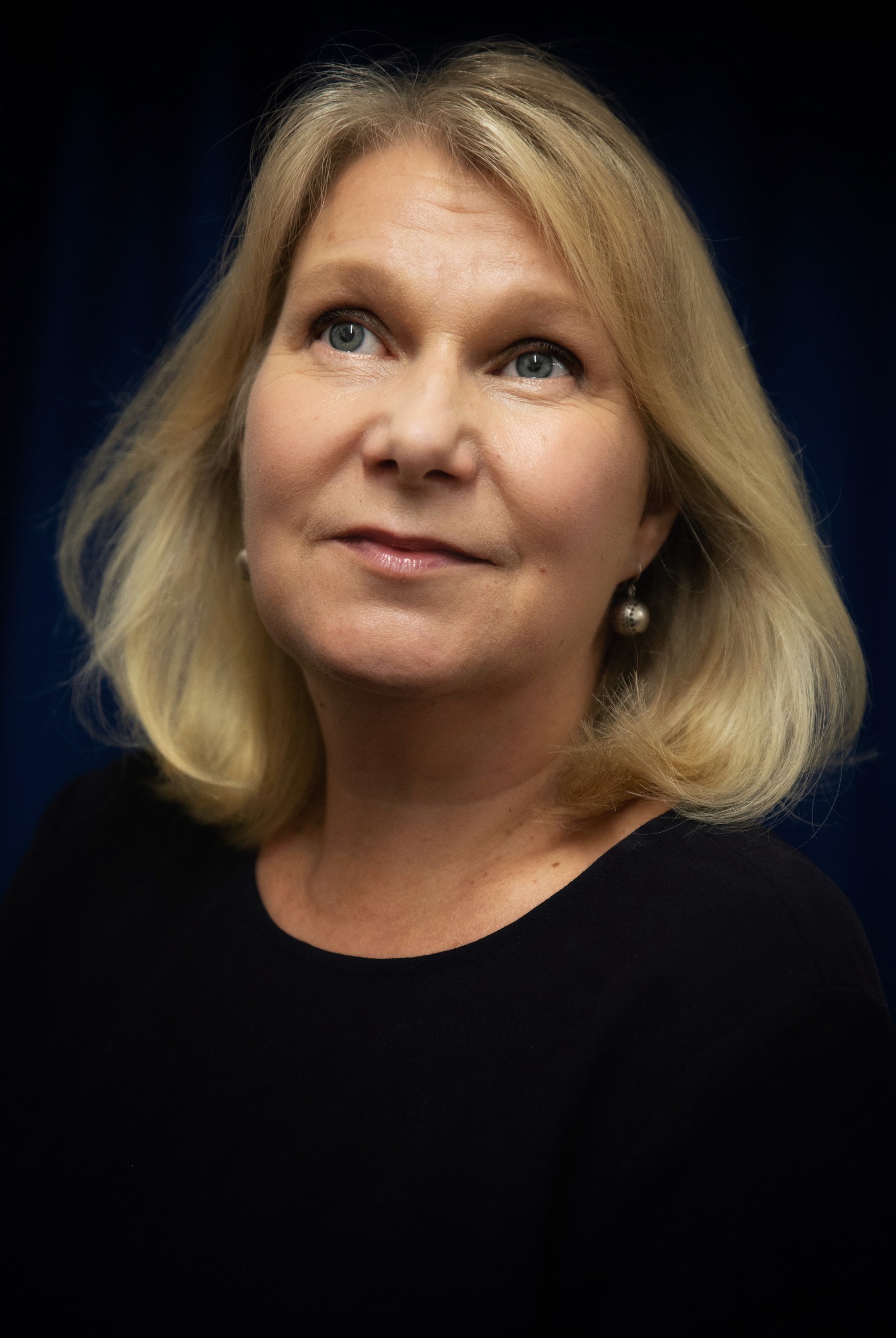 Muotokuva vaaleahiuksisesta naisesta, jolla on siniset silmät ja musta paita, tummansinisellä taustalla.